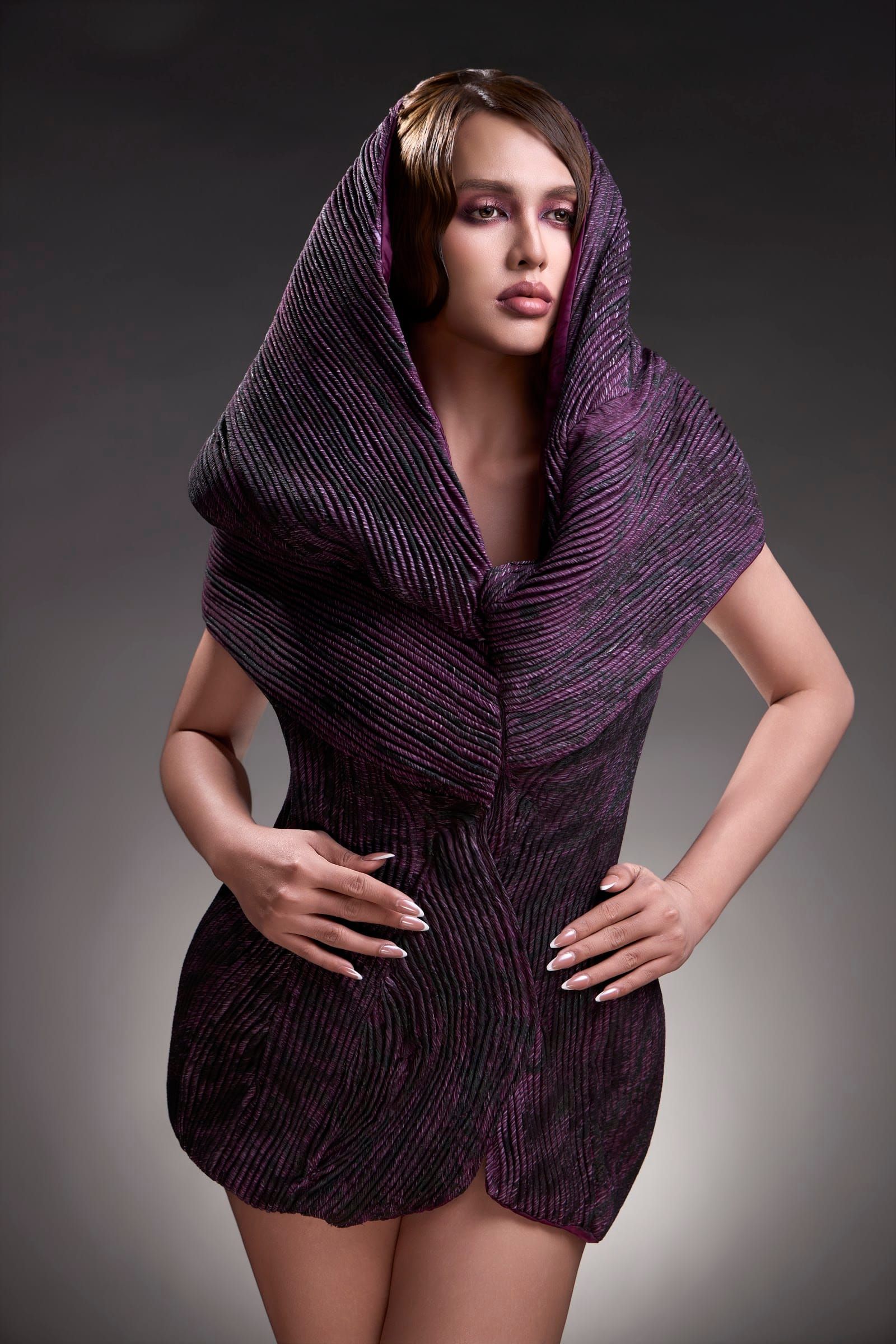 Full length of a woman in a purple pleated hood, hands on her hips, dark grey backdrop