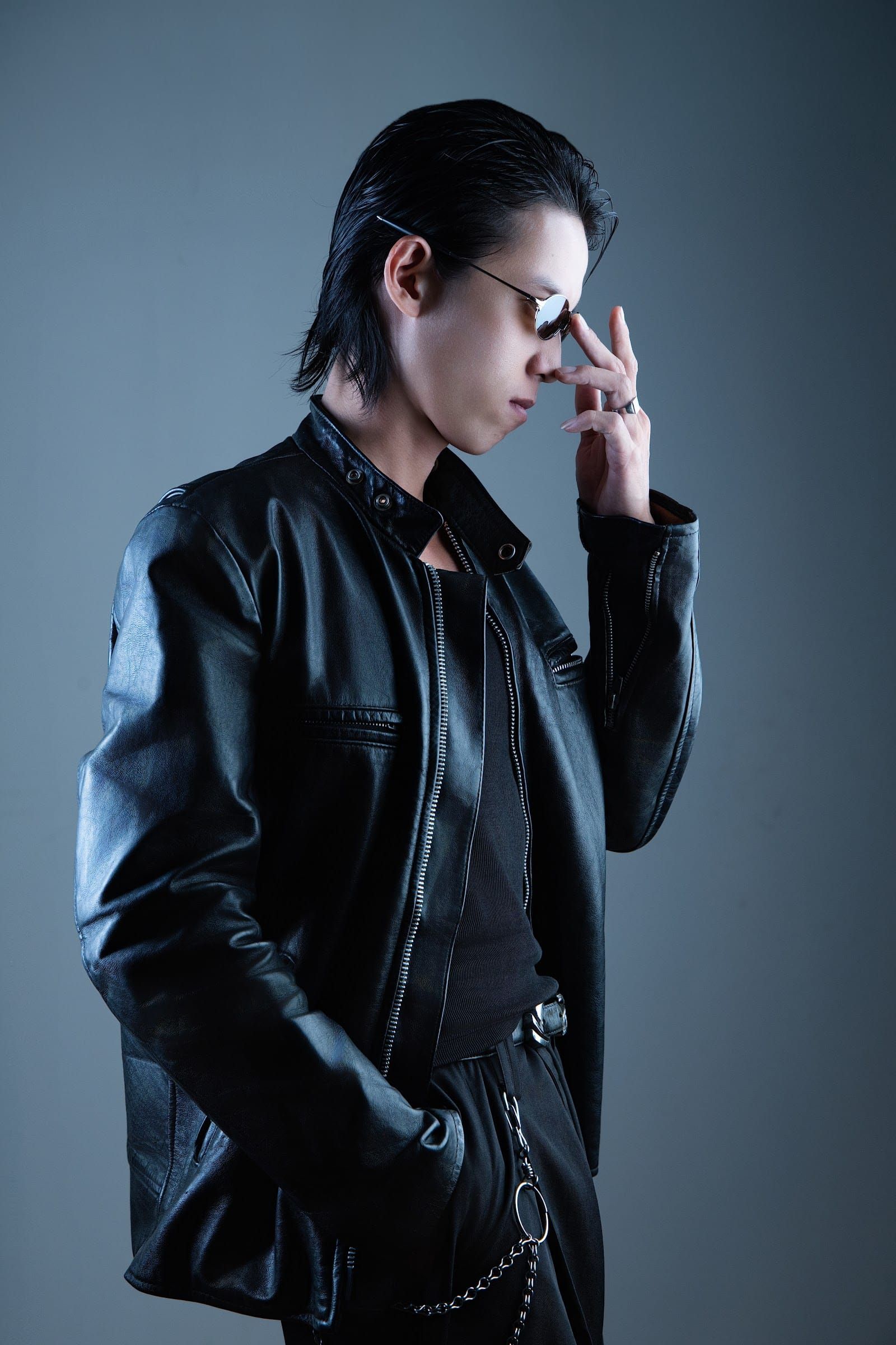 A model in a black leather jacket and sunglasses, one hand at her chin, blue-grey backdrop