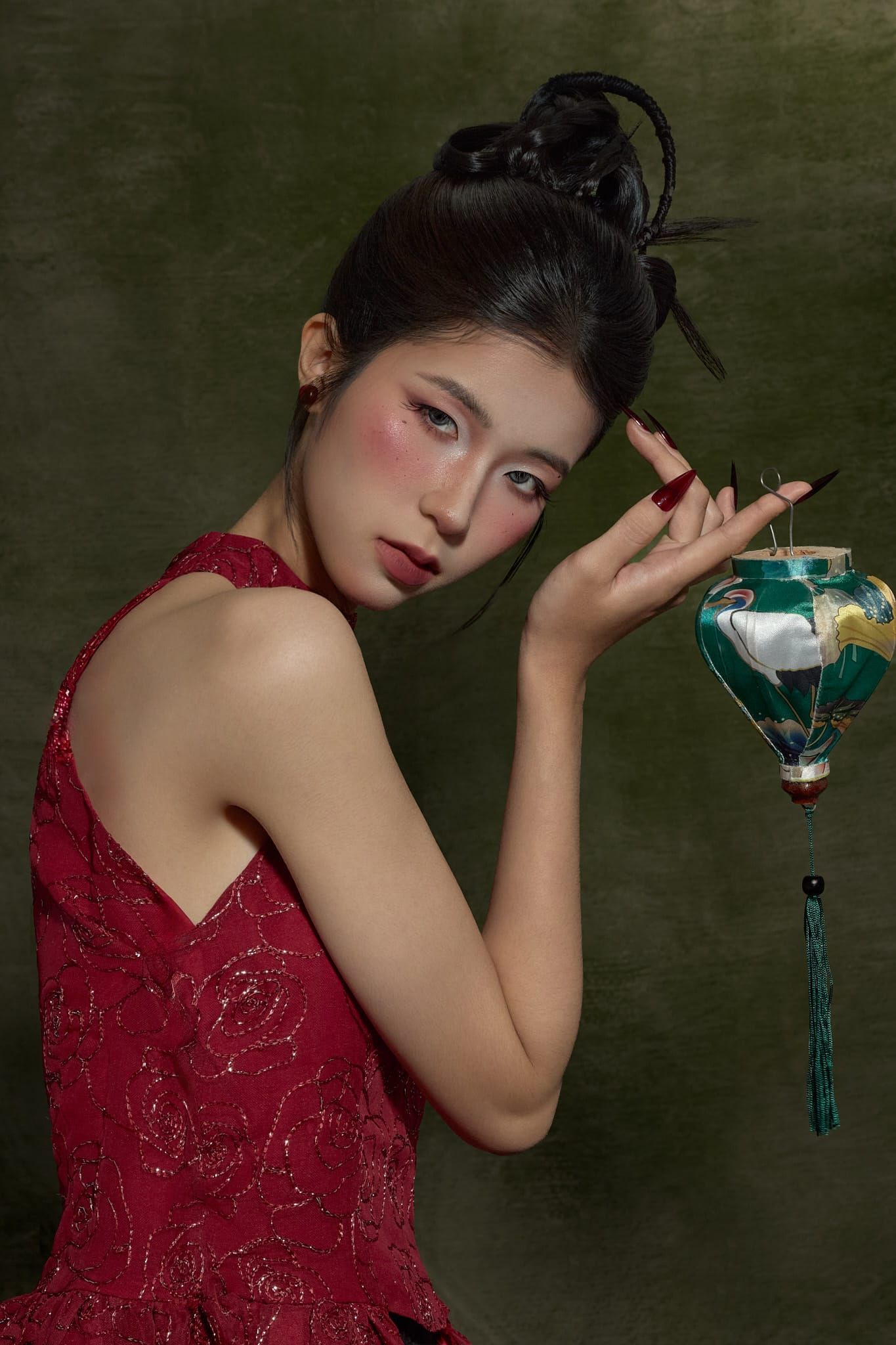 A woman in a red top holding a lantern, looking down, moss-green backdrop