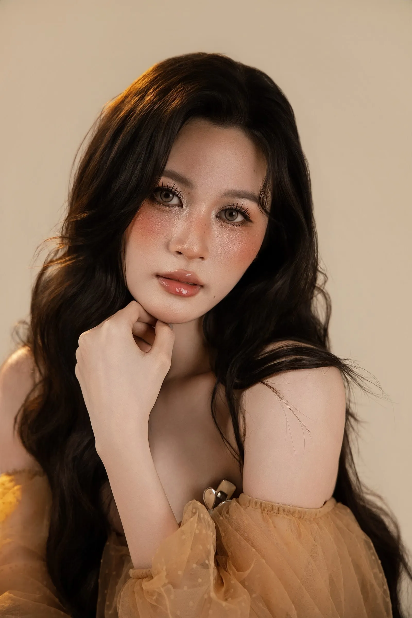 Warm-light portrait: a woman with long hair resting her hand on her cheek, a beige off-shoulder top