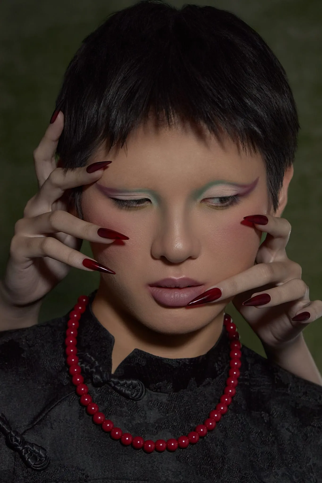 A face with graphic white makeup, both hands on the cheeks, a red bead necklace, moss-green backdrop